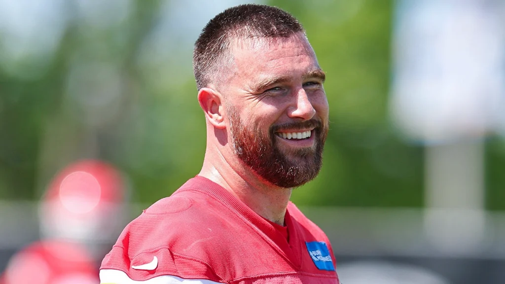 Smiling NFL player in a red practice jersey outdoors, featured in an article about Travis Kelce’s smile.